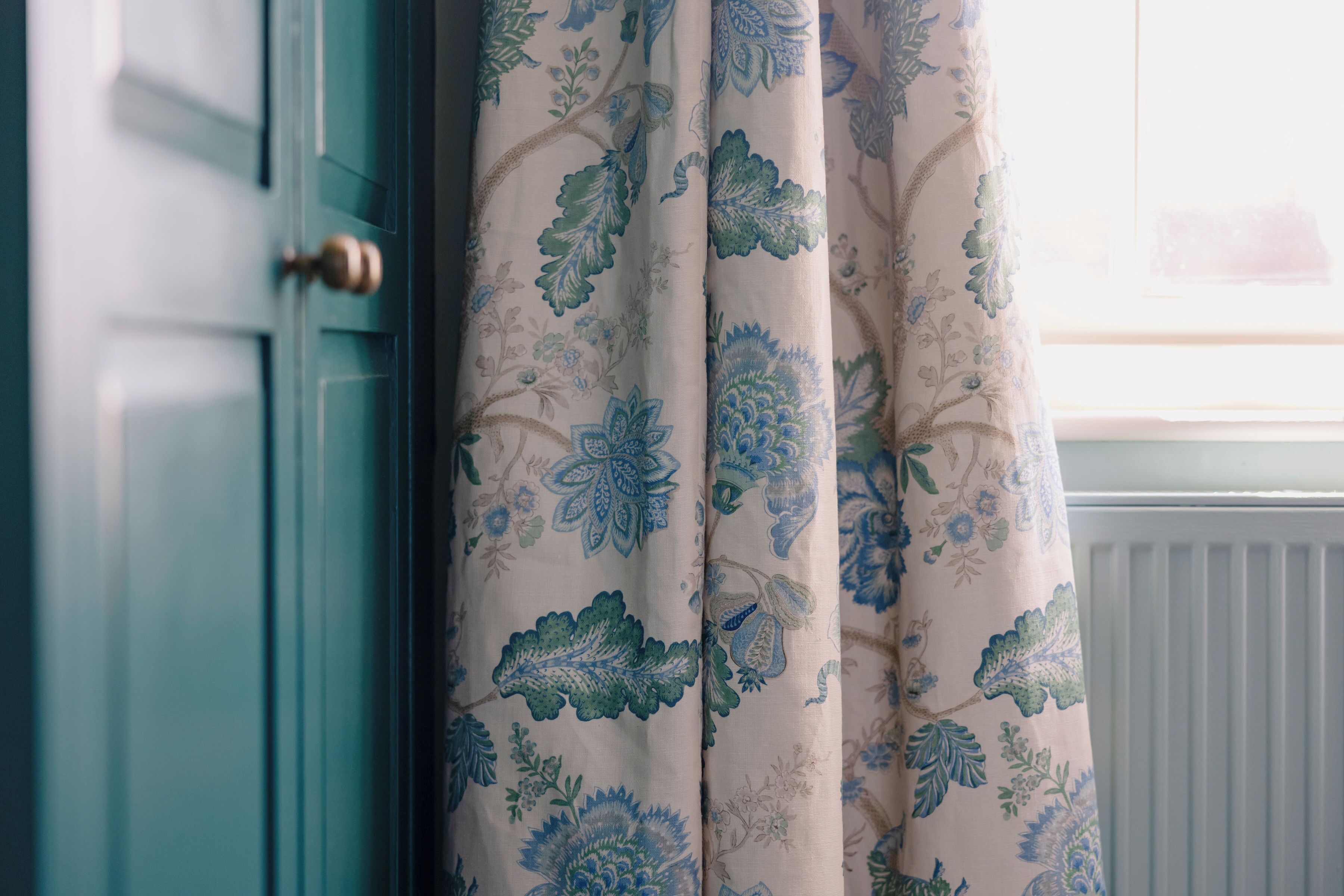 Eine blaue Tür mit Messinggriff, ein Fenster mit weißer Heizkörper und ein Vorhang mit Blumenmuster.