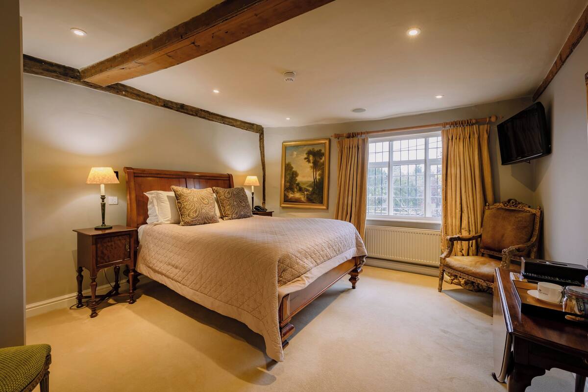 A bedroom with a large bed, a wooden headboard, a nightstand, a chair, a desk, a lamp, a painting, and a window with curtains.