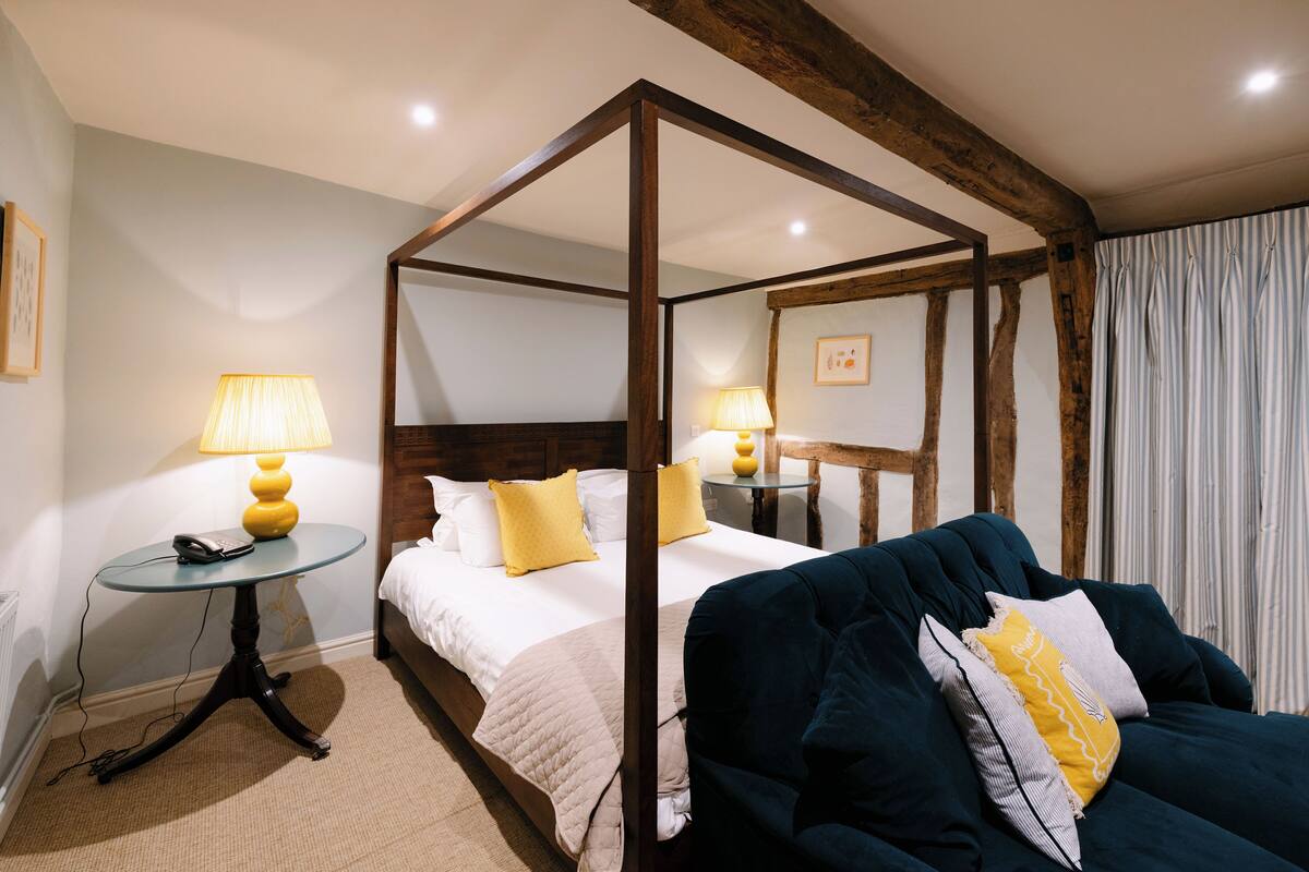A four-poster bed with a wooden frame, a bedside table with a lamp, a dark blue sofa with cushions, and a window with curtains.