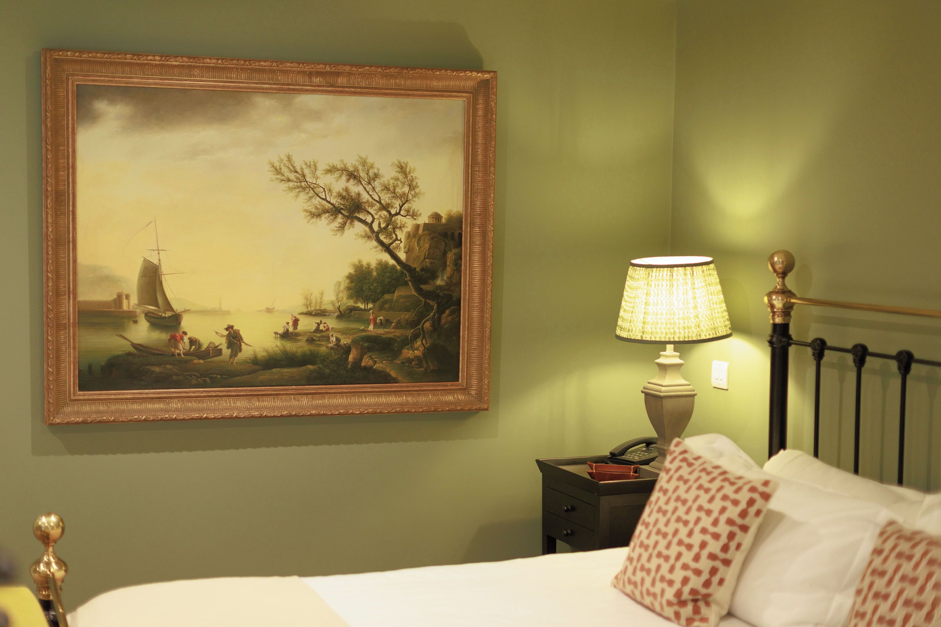 Un dormitorio con cama, almohadas, mesita de noche, lámpara y un cuadro en la pared.