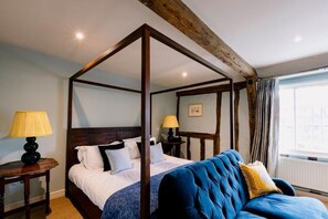 Uma cama de dossel com estrutura de madeira, roupa de cama branca e cabeceira estofada em azul. Há duas mesas de cabeceira com luminárias.