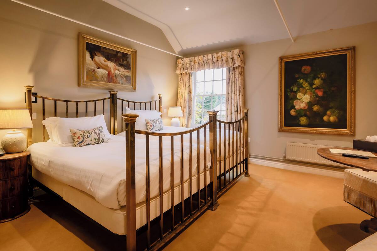 A bedroom with a four-poster bed, a nightstand with a lamp, a window with curtains, and a painting on the wall.