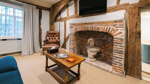 A room with exposed wooden beams, a brick fireplace, a leather armchair, a wooden coffee table, and a television mounted above the fireplace. - The Bildeston Crown (Bildeston)