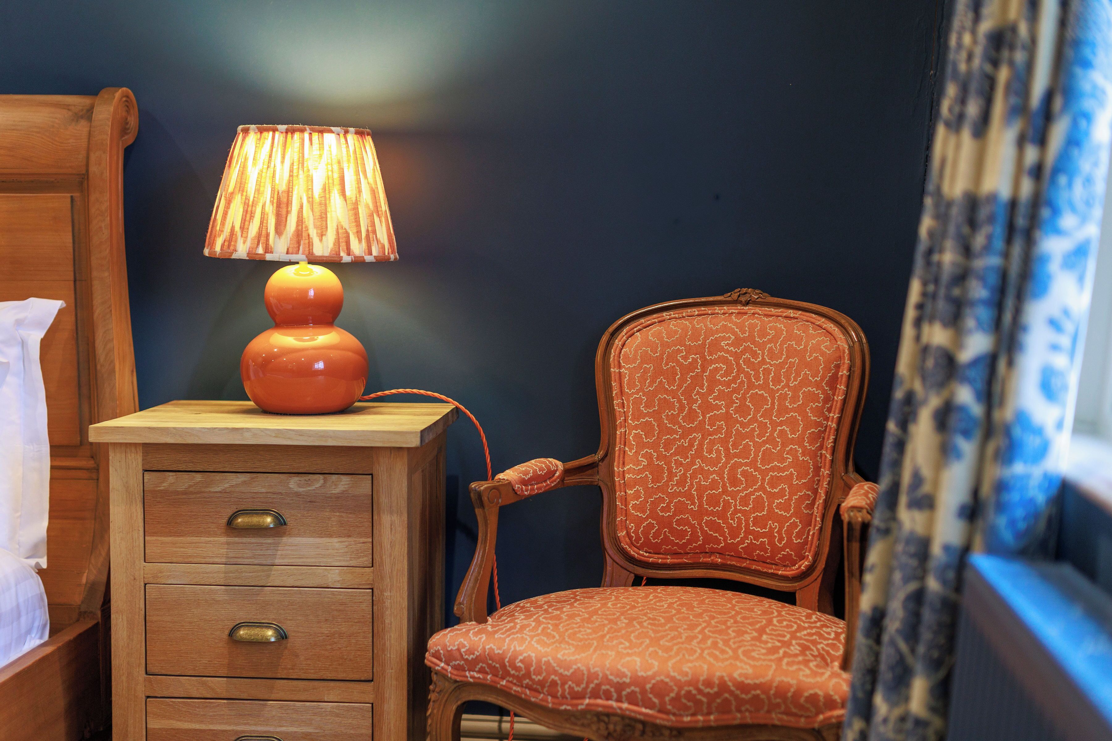 Une table de chevet en bois avec des tiroirs, une lampe et une chaise à motifs dans une pièce aux murs sombres.