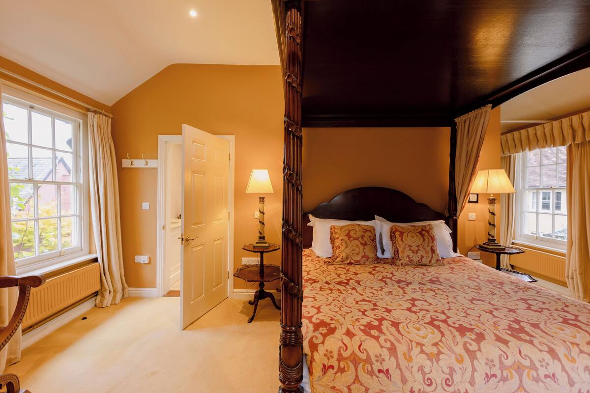 A bedroom with a four-poster bed, two bedside lamps, a radiator, and a window with curtains.