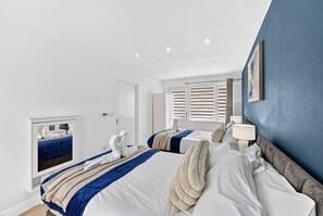 Ett hotellrum med två sängar, en blå accentvägg, ett fönster med persienner, en spegel och en sänglampa.