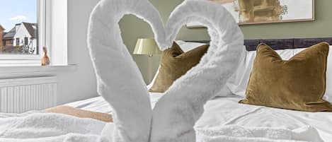Una cama con toallas blancas y mullidas dispuestas en forma de corazón, almohadas marrones y vistas a casas a través de una ventana.