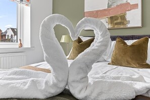 Ein Bett mit weißen, flauschigen Handtüchern, die zu einem Herz geformt sind, braunen Kissen und Blick auf Häuser durch ein Fenster.