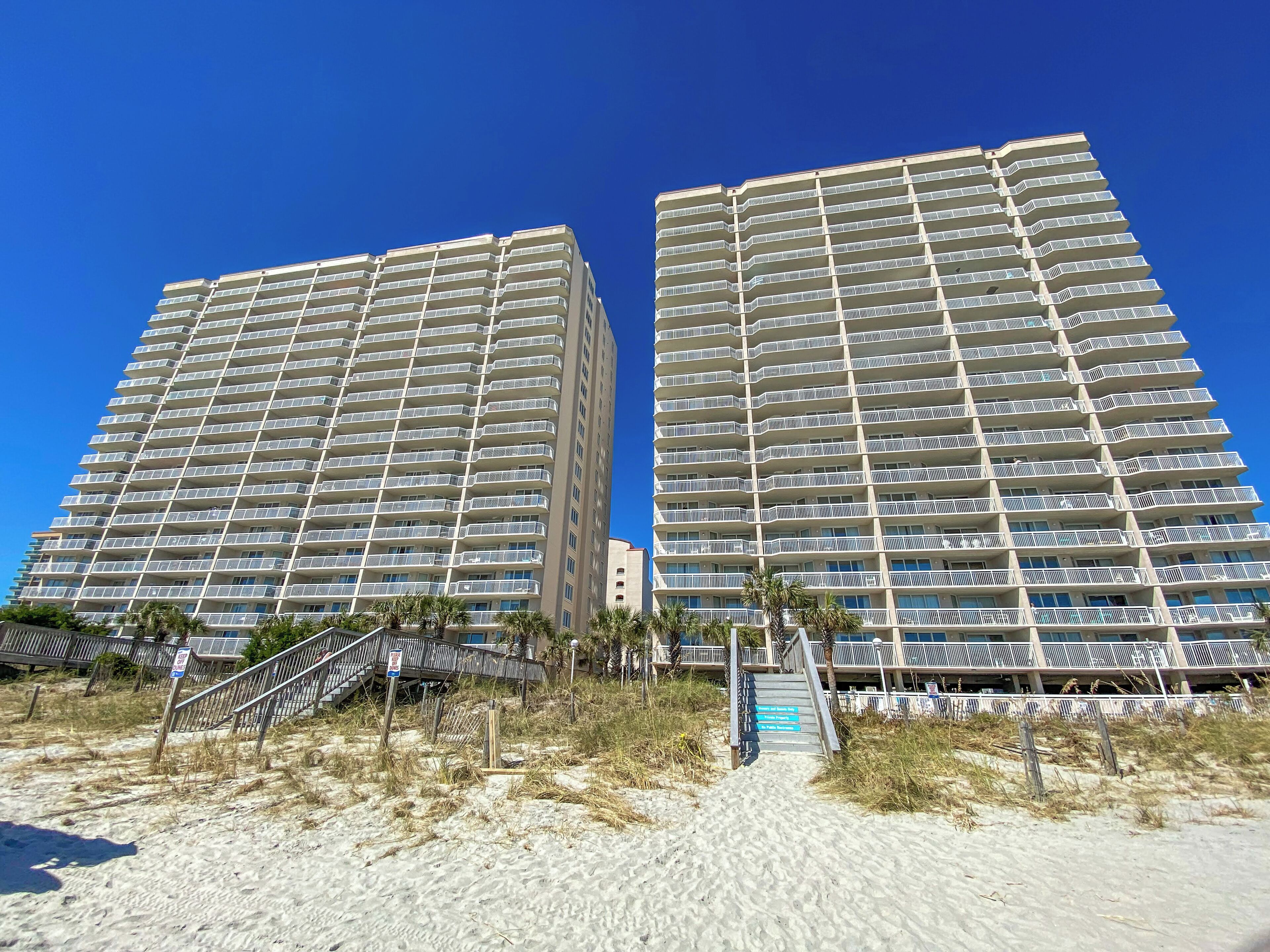 Two high-rise buildings with multiple balconies, a sandy beach area with a staircase leading to the buildings, and a clear blue sky.