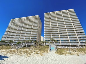 Deux immeubles de grande hauteur dotés de nombreux balcons, une plage de sable avec un escalier menant aux immeubles, et un ciel bleu azur.