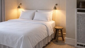 Een net opgemaakt bed met witte lakens, twee bedlampen, een klein houten krukje en een witte plafondlamp.