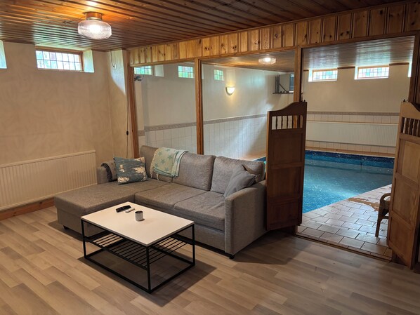 Ein Zimmer mit grauem Sofa, einem kleinen Tisch, einer Holztür und Blick auf einen Swimmingpool.