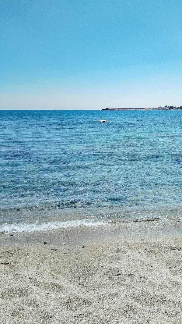 A beach with clear water, pebbly shore, and distant buildings. - Loving Gift - Giardini Naxos (Giardini Naxos)