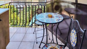 A balcony with a tiled floor, a round table with a patterned tablecloth, and a chair with a decorative backrest. - Loving Gift - Giardini Naxos (Giardini Naxos)