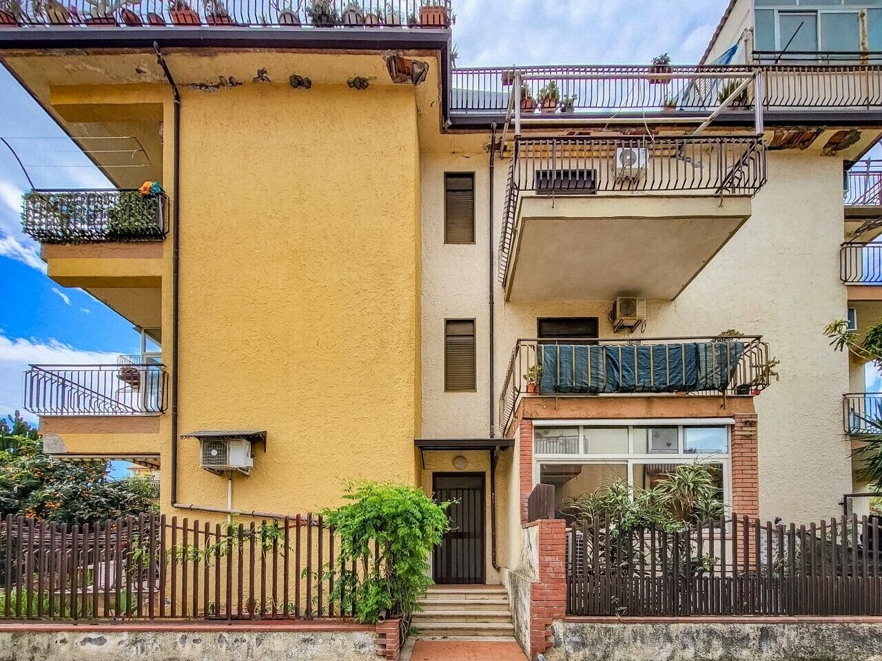 A two-story residential building with balconies, a visible front door, and a small garden area.