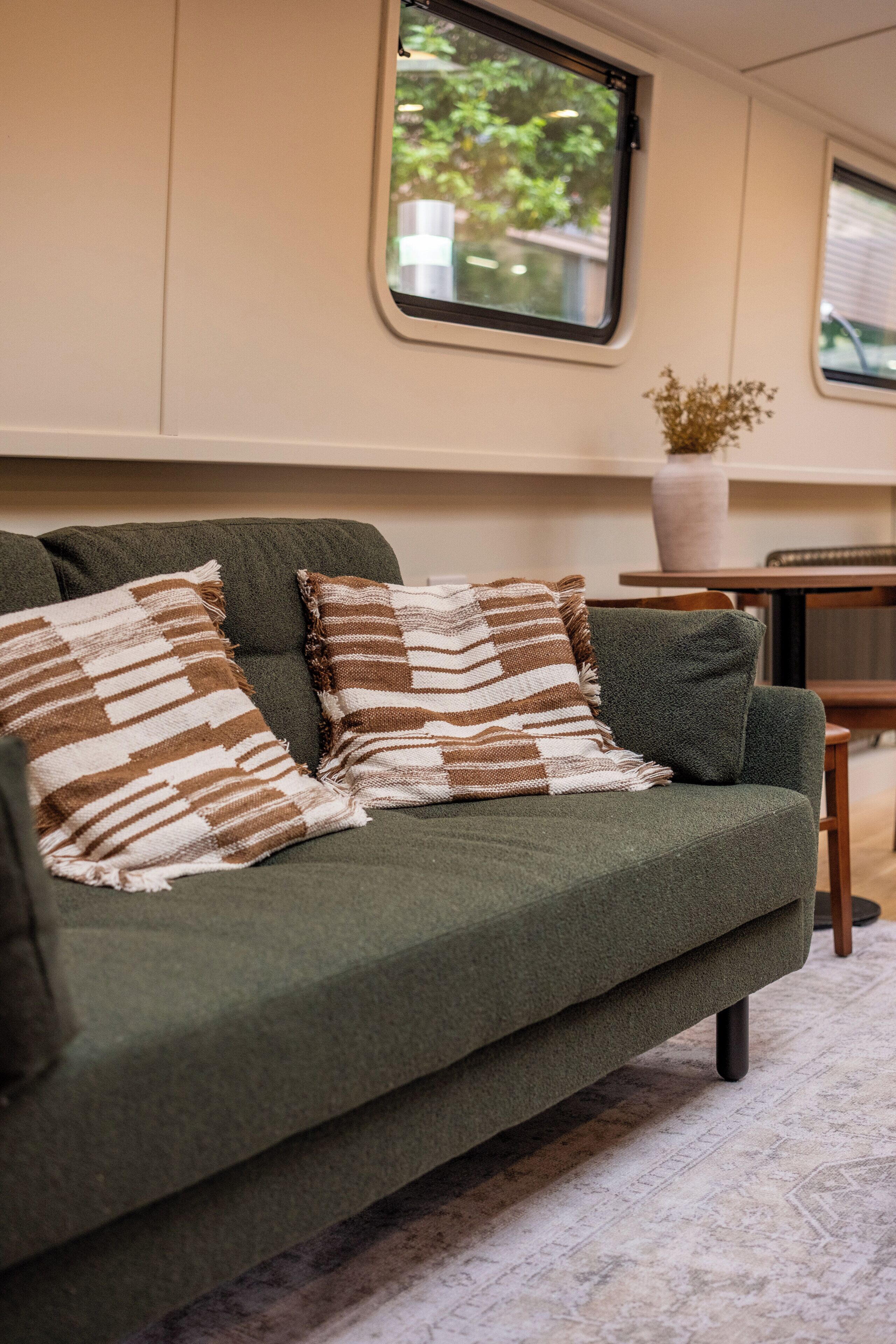 A green sofa with striped pillows, a vase with dried flowers, and a window with a view of greenery.