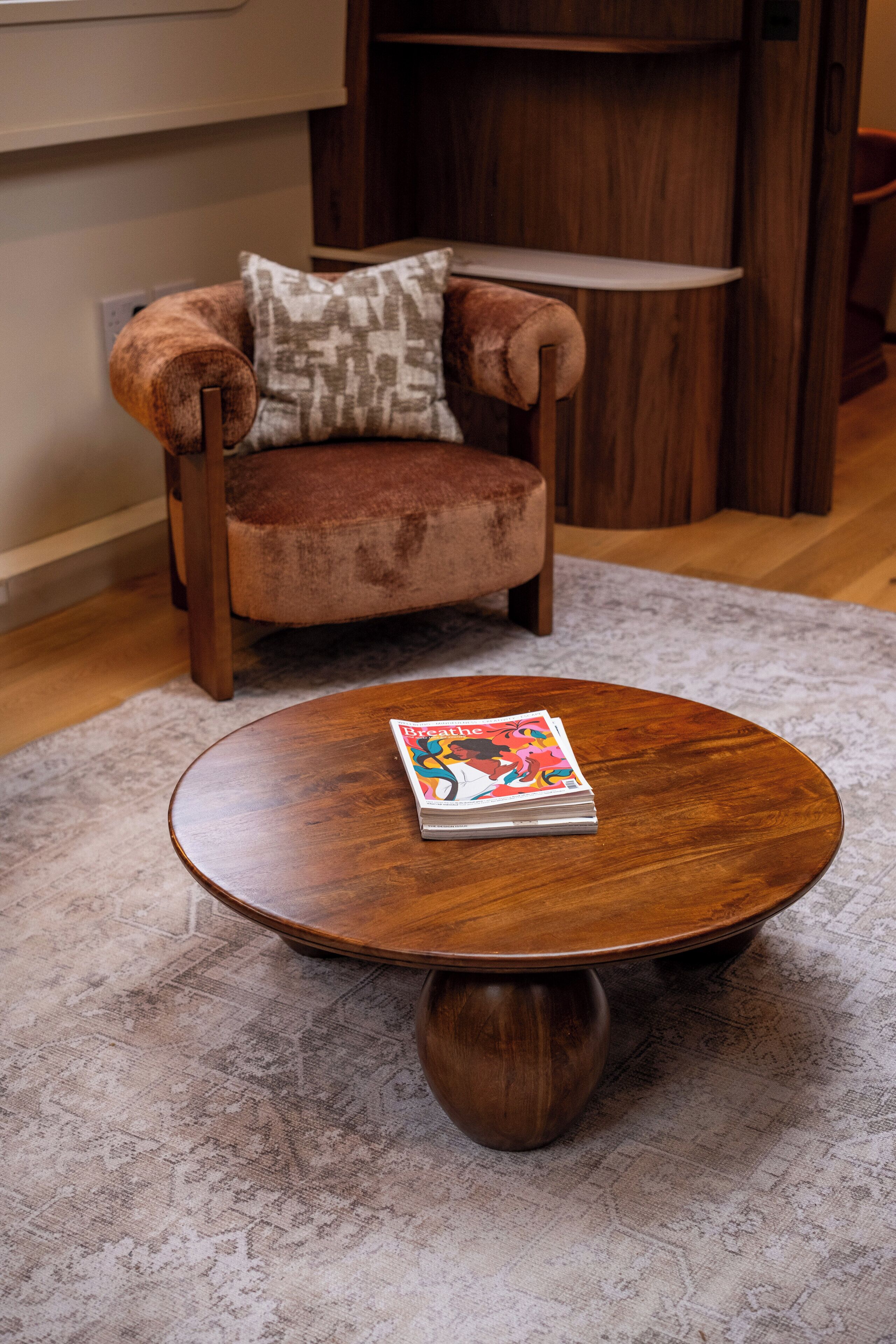 A round wooden coffee table with a magazine on it, placed on a patterned rug.