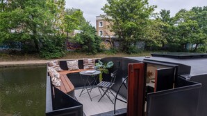 A modern outdoor seating area with a table and chairs, a small garden, and a view of the river. - Gainsborough Boat House (London)
