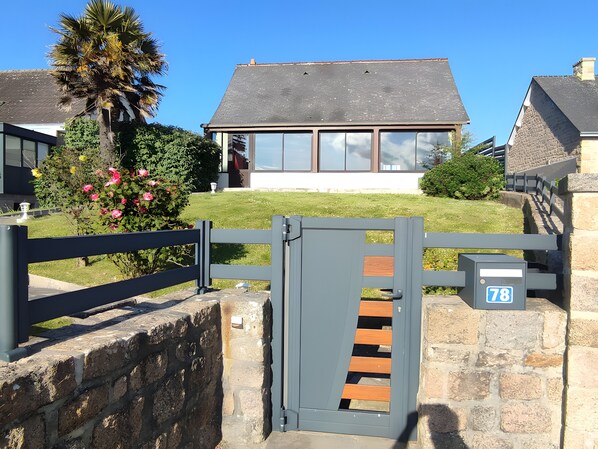 Ein verschlossenes Tor mit einer Steinmauer, ein Haus mit grauem Dach und einem strahlend blauen Himmel.