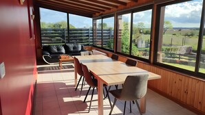A modern dining area with a wooden table and chairs, a sofa, and large windows offering a view of the outdoors. - Holiday Home “Gîte for 10 in Barfleur” with Private Terrace, Garden & Wi-Fi (Barfleur)