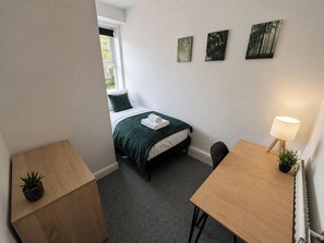 Una habitación pequeña y ordenada con una cama, un escritorio, una silla y una lámpara. Hay tres cuadros en la pared.