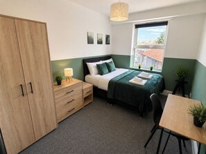 A bedroom with a bed, bedside table, lamp, wardrobe, window, and a small table with a chair.