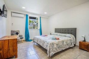 Ein Schlafzimmer mit einem Bett, einem Schreibtisch, einem Stuhl, einem Fenster mit Vorhängen und einer Klimaanlage.