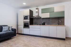 Eine moderne Küche mit integriertem Backofen, Mikrowelle und Herd, ein graues Sofa mit gemusterten Kissen und ein Fliesenboden.