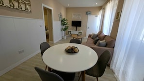 Un comedor con una mesa redonda, sillas y un tazón con objetos decorativos. Se observa un sofá y un televisor al fondo.