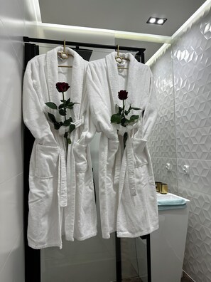 Deux peignoirs blancs ornés de roses sont suspendus à un porte-peignoir dans une salle de bain dont le mur est recouvert de carreaux géométriques.