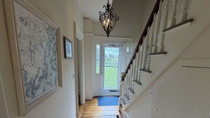 Un pasillo con piso de madera, un candelabro, una escalera con barandillas blancas y una puerta que da al exterior.