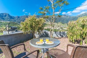 Uma mesa com dois pratos de comida e dois copos de suco de laranja, rodeada por cadeiras de vime, com vista para uma montanha ao fundo.