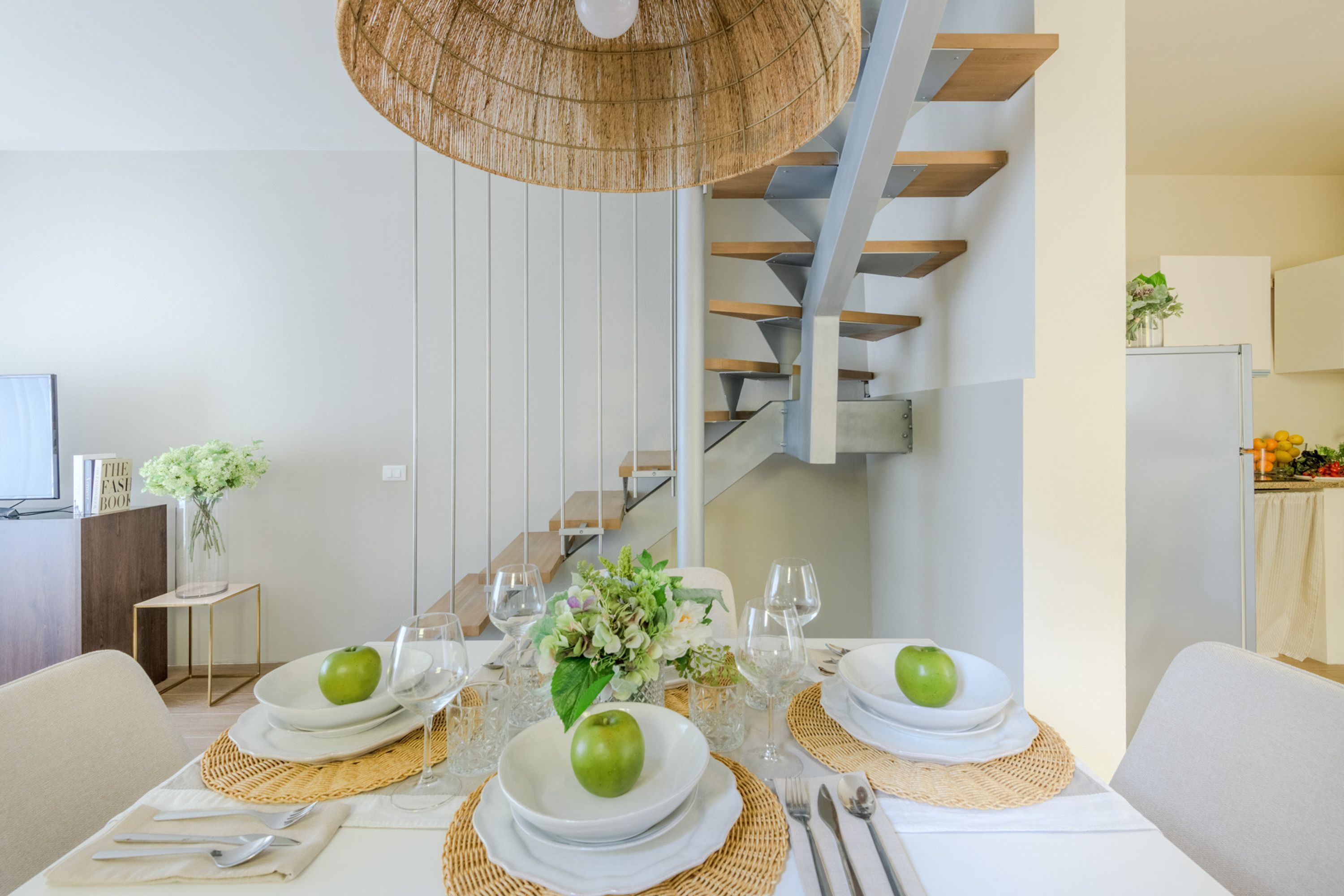 Zona pranzo con tavolo apparecchiato per quattro, una scala e una cucina sullo sfondo.