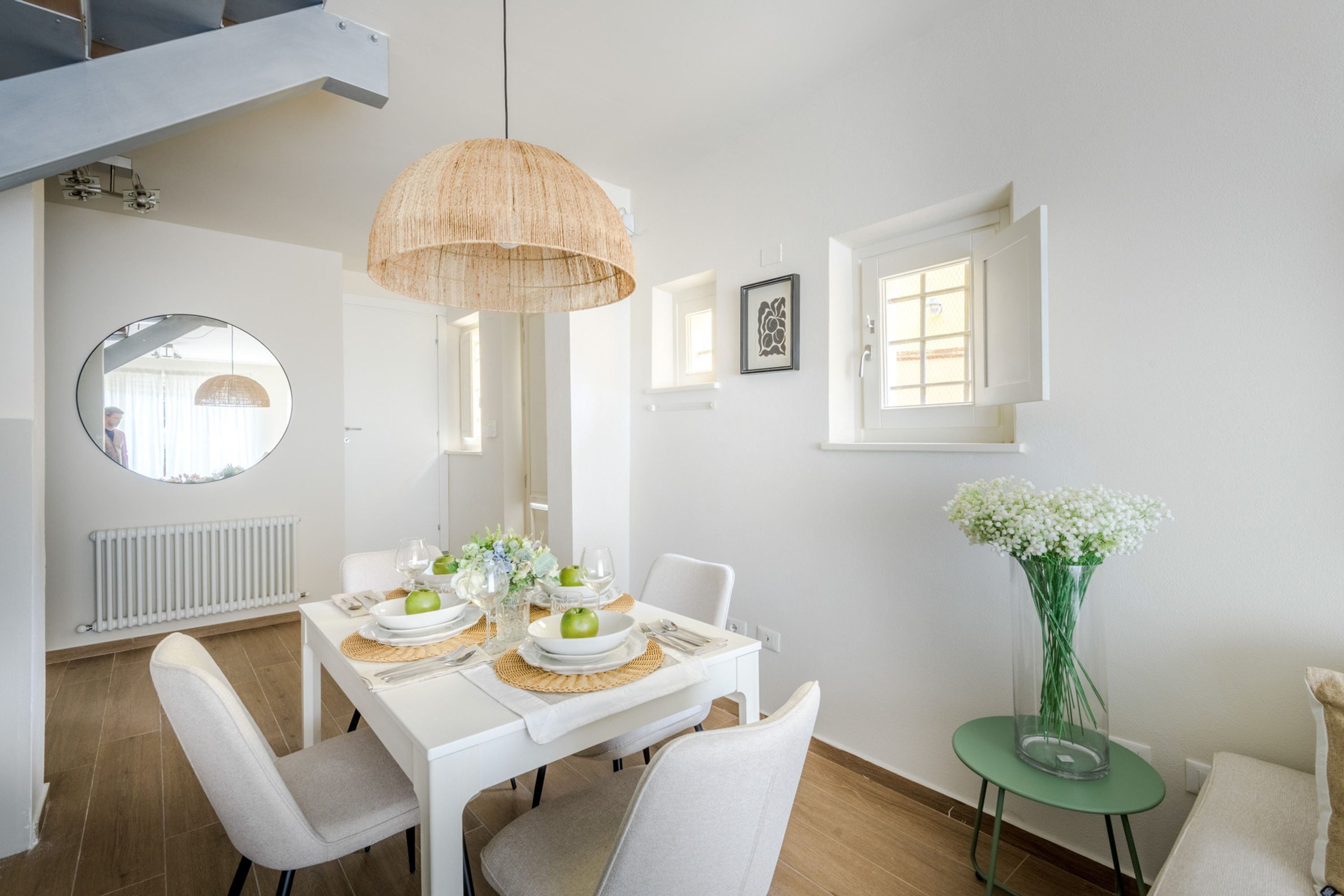 A dining area with a white table set for two, a hanging light fixture, a round mirror, and a window with a view.