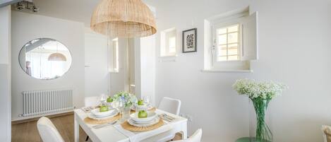 A dining area with a white table set for two, a hanging light fixture, a round mirror, and a window with a view.