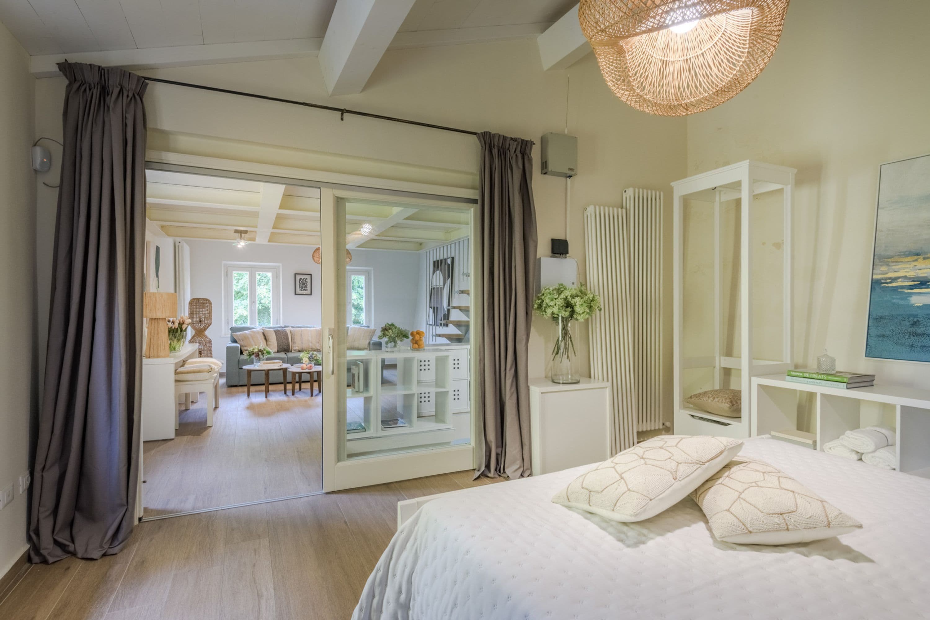 Una camera da letto con un letto, una grande finestra, un armadio bianco e una porta vetrata scorrevole che dà su un soggiorno.