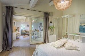 Ein Schlafzimmer mit Bett, einem großen Fenster, einem weißen Kleiderschrank und einer Glastür, die in einen Wohnbereich führt.