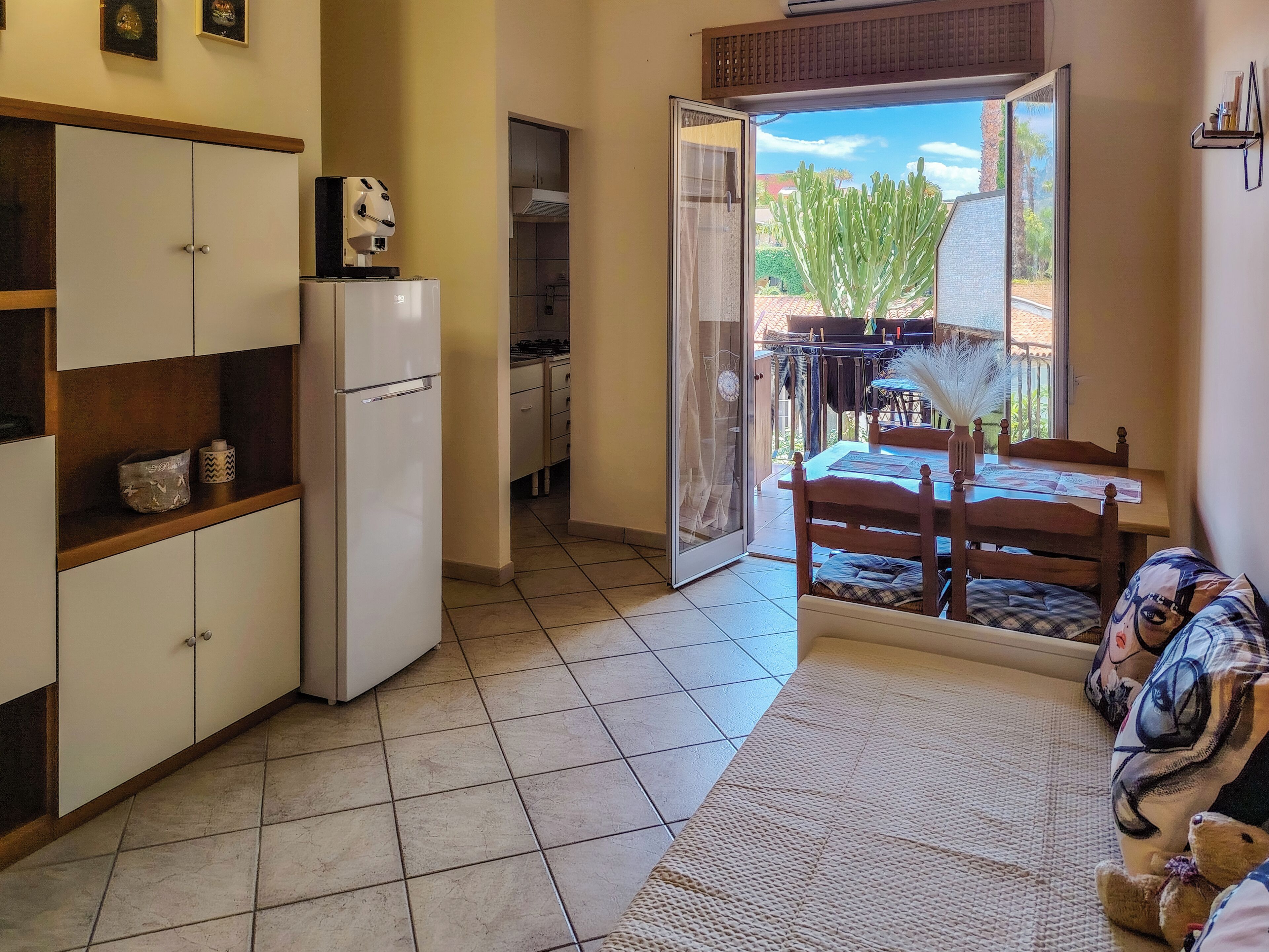 Uma cozinha compacta com geladeira, uma área de refeições com mesa e cadeiras, e uma porta de vidro deslizante que dá para uma varanda com um cacto.