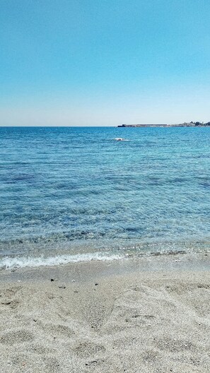Une plage à l’eau d’un bleu limpide, avec un rivage de galets et une côte lointaine ponctuée de bâtiments.