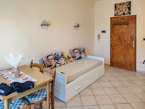A small, neatly arranged room with a bed, a table with a chair, a wooden door, and a wall-mounted shelf. - Loving Gift (Giardini Naxos)