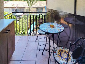 Un balcon doté d’un sol carrelé, d’une table recouverte d’une nappe aux motifs colorés, d’une chaise et d’une rambarde.