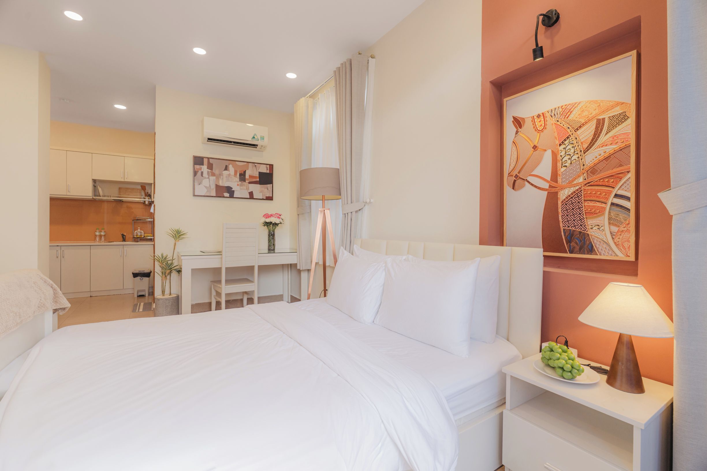 A hotel room with a bed, bedside table with a lamp, a desk with a chair, a kitchen area, and a wall-mounted air conditioner.
