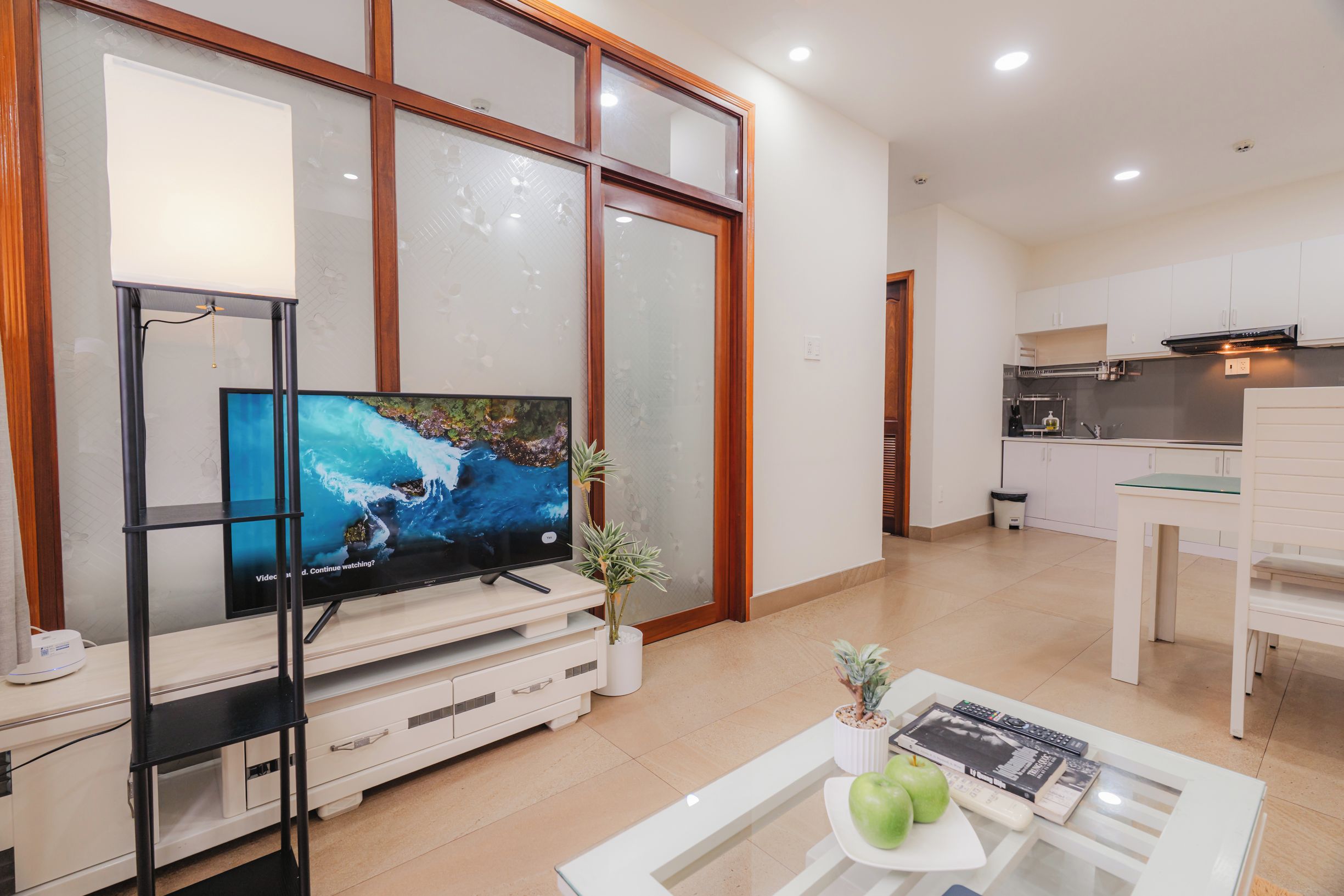 Uma sala de estar moderna com televisão de tela plana, uma mesa de centro com livros e uma planta, e uma cozinha ao fundo.