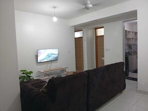 Ein Wohnzimmer mit einem dunkelbraunen Sofa, einem flachbildfernseher an der Wand, einem kleinen weißen Tisch und einer Topfpflanze.