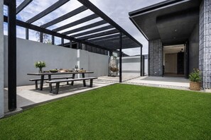 A modern outdoor patio with a wooden table set for dining, a grassy lawn, and a covered seating area. - Stackbrae House with Mountain Views (Wānaka)