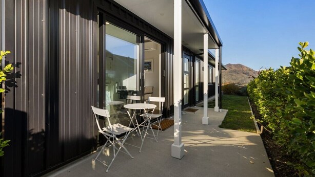 A modern building with large glass windows, a covered outdoor seating area with white chairs, and a view of a mountain in the background.