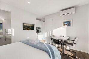 A modern hotel room with a bed, a desk, and a chair, a small kitchenette, and an air conditioning unit.