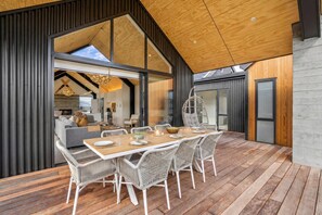 A modern interior with a dining area, white wicker chairs, a wooden table, and a large window. - Architectural Northlake Retreat | Sleeps 8, Wood-Burning Fireplace, Media Room & Covered Outdoor Deck (Wānaka)