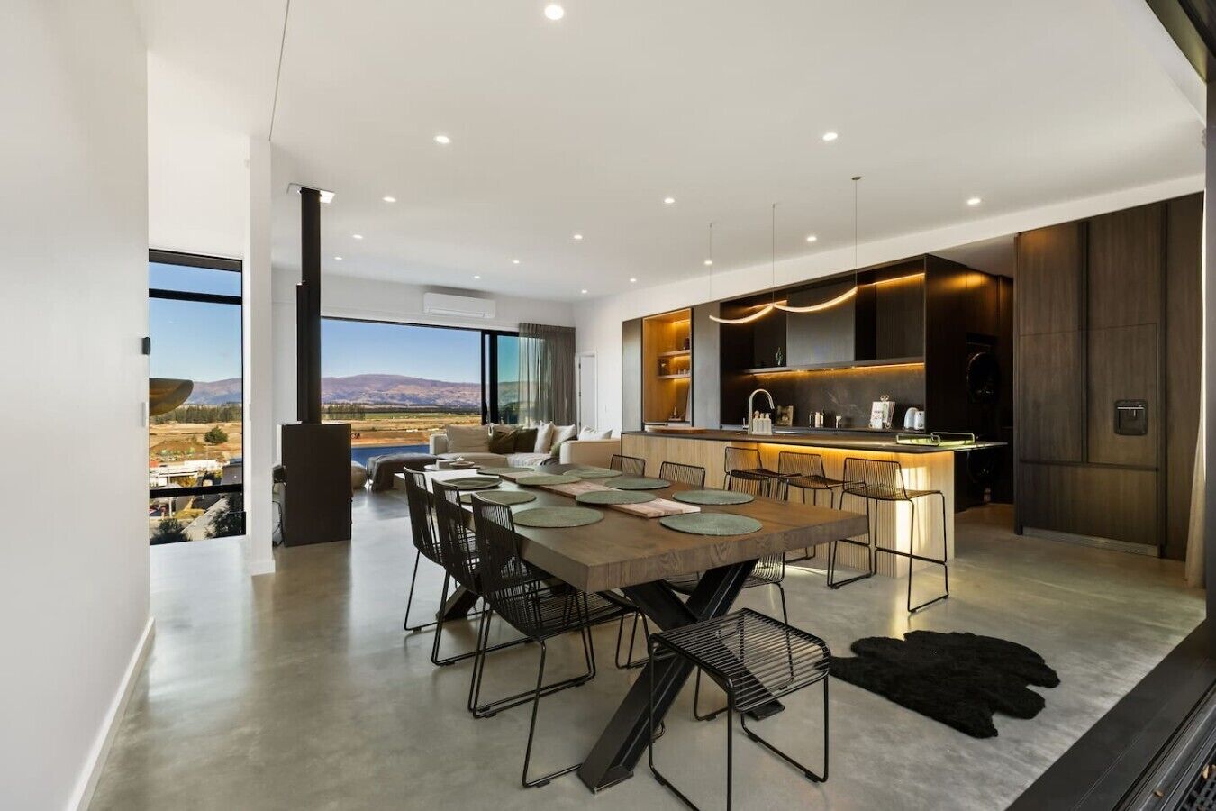 A modern kitchen with a central island, a dining area with a large table and chairs, and a view of a mountainous landscape through large windows.