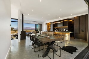 Eine moderne Küche mit einer zentralen Kochinsel, ein Essbereich mit großem Tisch und Stühlen sowie ein Blick auf eine gebirgige Landschaft durch große Fenster.